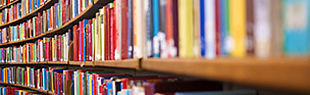 Das Bild zeigt ein Regal einer Bibliothek gefüllt mit Büchern. Vorn, auf der rechten Seite etwas unscharf, nach hinten auf der linken Seite deutlicher werdend mit fünf Regalböden.