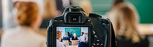 Auf dem Bild ist eine Videokamera abgebildet, die hinten in einem Klassenraum steht und das Geschehen im Klassenraum aufzeichnet. 