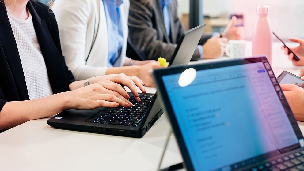 Auf dem Bild sind mehrere Laptops abgebildet sowie Personen, die an den Laptops arbeiten. 